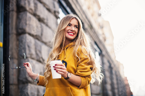 Stylish happy young woman wearing boyfrend jeans, white sneakers bright yellow sweetshot.She holds coffee to go. portrait of smiling girl in sunglasses and with bag photo