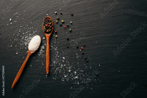 Sea salt and mixed peppercorn in wooden spoons. Top view on black slate board with copy space.