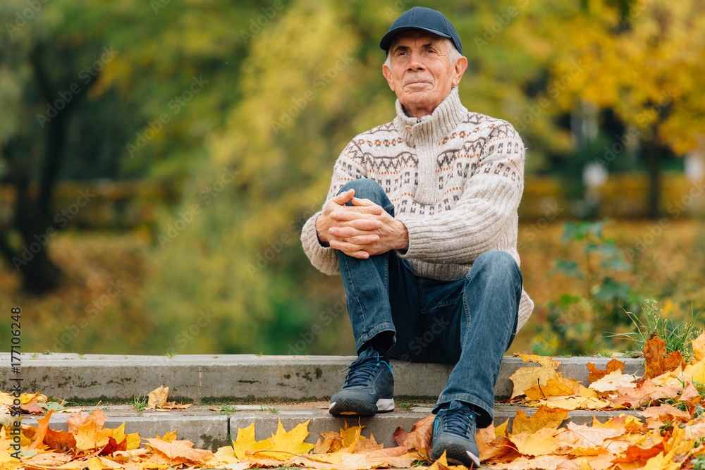 Portrait of pretty senior man in cap sitting in colorful autumn park. Pensioner resting outdoors. Beutiful fall.