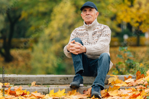 Obraz na płótnie Portrait of pretty senior man in cap sitting in colorful autumn park. Pensioner resting outdoors. Beutiful fall. w salonie