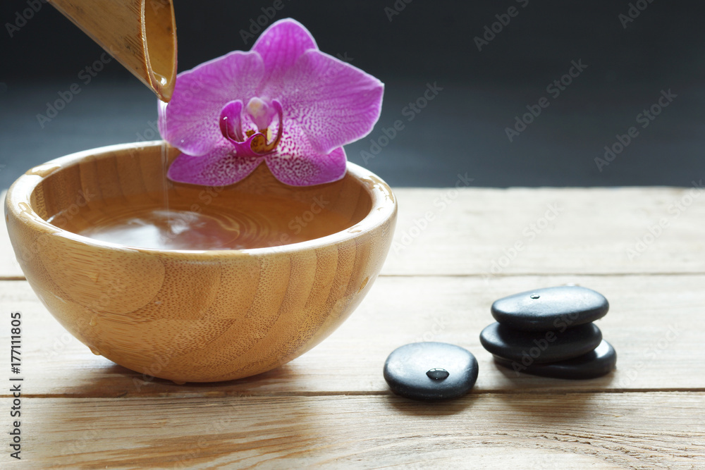 Bowl, which pours transparent water from the bamboo, orchid flowers and stones for a hot massage on a wooden table Bowl, which pours transparent water from the bamboo, orchid flowers and stones for a hot massage on a wooden table