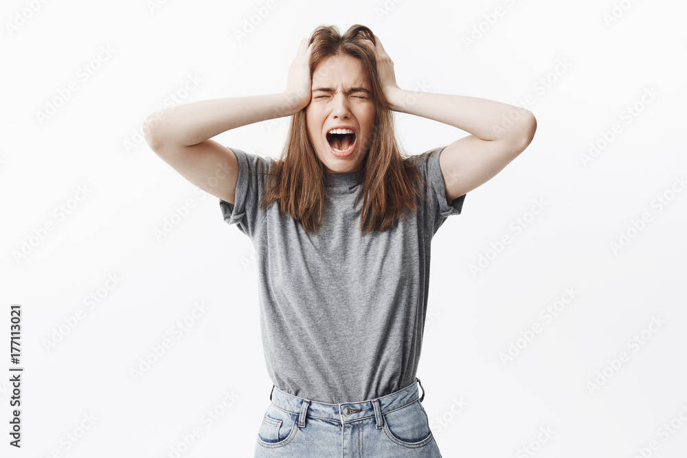Young beautiful european student with dark hair in grey t-shirt and jeans holding head with hands, screaming with closed eyes,after being attacked with big amount of work Young beautiful european student with dark hair in grey t-shirt and jeans holding head with hands, screaming with closed eyes,after being attacked with big amount of work
