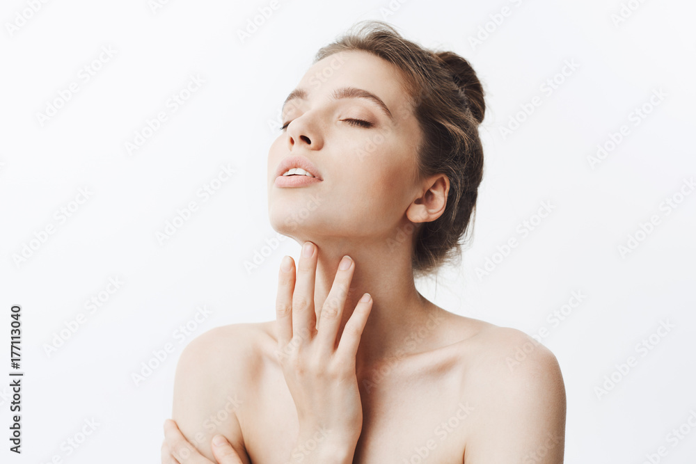 Young beautiful dark-haired caucasian woman with bun hairstyle and naked shoulders raiding head with closed eyes touching neck. Girl using body cream after bath. Young beautiful dark-haired caucasian woman with bun hairstyle and naked shoulders raiding head with closed eyes touching neck. Girl using body cream after bath.