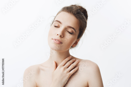 Portrait of sleepy good-looking young student girl with dark hair in bun hairstyle being naked, touching shoulders with hand and closed eyes, taking shower after long day at university.