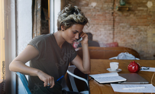 Young blonde woman is talking with friends by a mobile phone while sitting in a coffee shop. Attractive female is making calls by a smartphone while sitting with a portable computer.