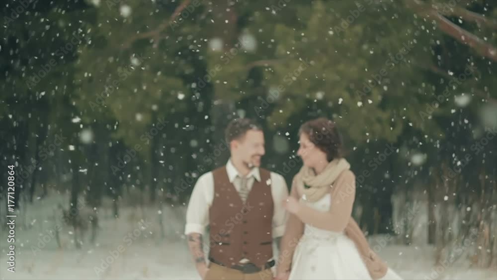 blurred loving couple walking in winter christmas snowfall and smiling. Rustic Style. Lovely Girl in Long Wedding White Dress Smiling, Green Pine Forest on Background. Brown Style Wedding. Slow Motion blurred loving couple walking in winter christmas snowfall and smiling. Rustic Style. Lovely Girl in Long Wedding White Dress Smiling, Green Pine Forest on Background. Brown Style Wedding. Slow Motion