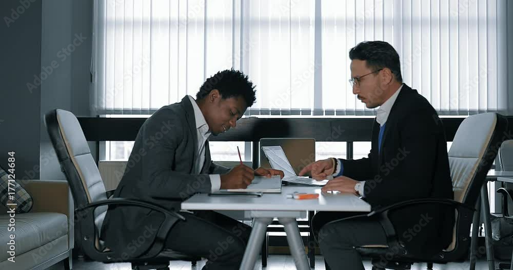 two young businessman talking at desk in office. European and African businessmen two young businessman talking at desk in office. European and African businessmen