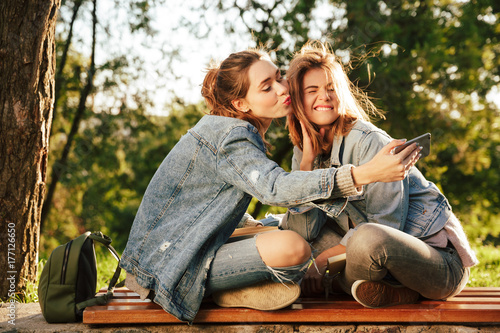 Close-up photo of two brunette teen girl having fun while taking selfie on smartphone in park outdoor