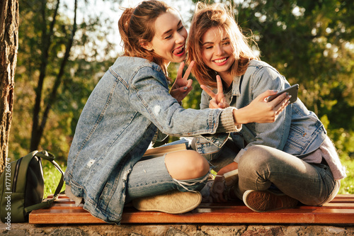 Two cheerful female friends showing peace sign wile taking selfe on smartphone in sunny park