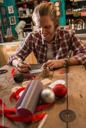 Young adult man with long blonde hair writing christmas card seated on wooden table in cozy apartment indoor.