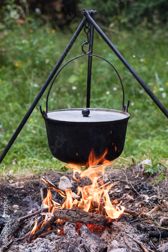 The cooking pot with lid which is hanging on the tripod above campfire