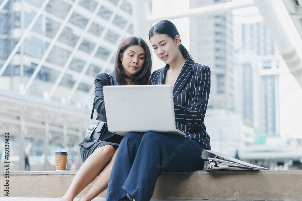 Two beautiful asian business women are are working on laptop with document to prepare for meet customer while sitting on stair outside office Two beautiful asian business women are are working on laptop with document to prepare for meet customer while sitting on stair outside office