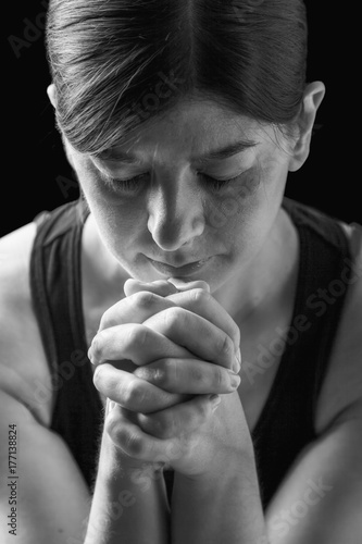Low key portrait of a faithful woman praying, hands folded in worship to god, head down and eyes closed in religious fervor, on a black background. Concept for religion, faith, prayer and spirituality