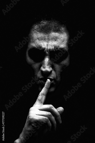 Angry mature man with an aggressive look making the silence sign in a threatening and creepy way. Black and White, black background. Concept for secret, threat, anger, rage, violence, danger, menace.