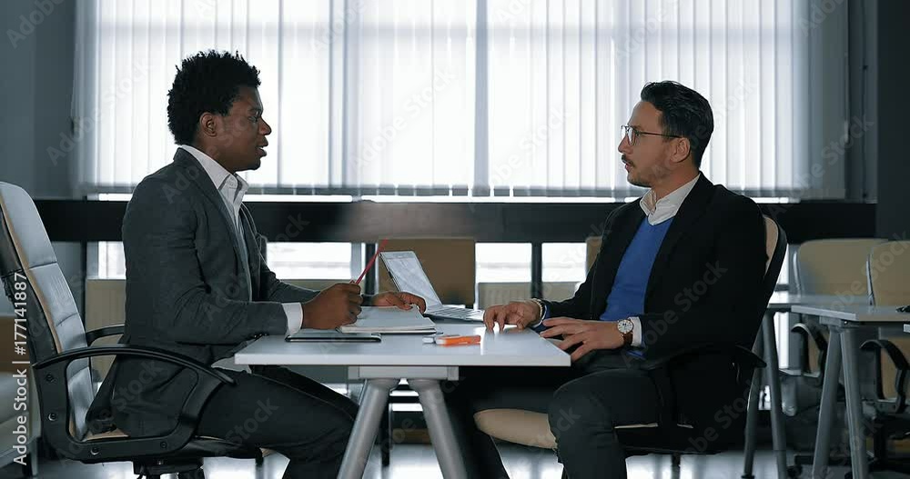 two young businessman talking at desk in office. European and African businessmen two young businessman talking at desk in office. European and African businessmen