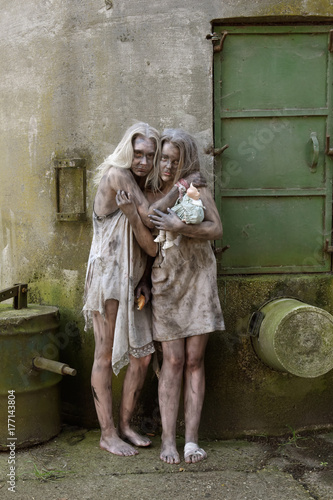 A scene of poverty with two young homeless girls.  They are seen to be situated in a poor unclean environment wearing rags as clothing. Their faces reveals sadness and sorrow in them.