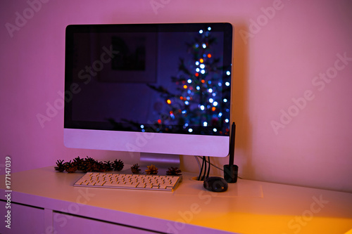 Christmas workspace interior with decorated christmas tree reflection in monitor and pine cones on the table. Bokeh lights. photo