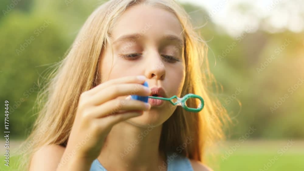 Portrait of a long-haired teen girl blowing soap bubbles. Then - smiling and looking into the camera. Park in the background. Close up Portrait of a long-haired teen girl blowing soap bubbles. Then - smiling and looking into the camera. Park in the background. Close up