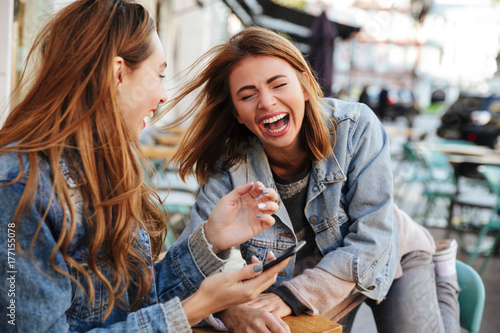 Two overjoyed brunette woman in jeans wear laughing while sitting at city cafe