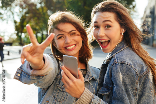 Two happy teen girl showing peace sign while listening to music on smartphone, looking at camera, outdoor