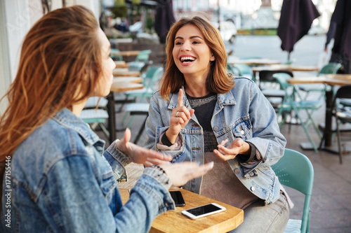 Billede på lærred Two young cheerful female friends talking while sitting in cite cafe