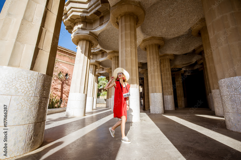 Fototapeta premium Happy woman tourist in red dress with hat walking near the columns visiting famous Guell park in Barcelona