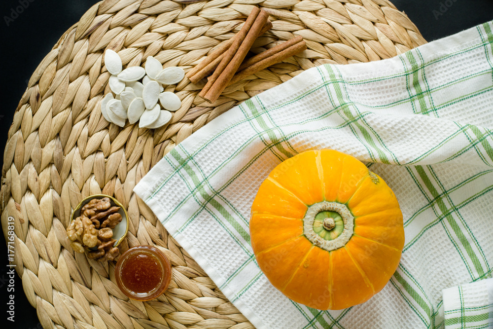 Small pumpkin with seeds,  little glass can of honey, walnuts and cinnamon sticks on a circle mat/napkin made of water hyacinth on a black background and green-white checkered waffle towel