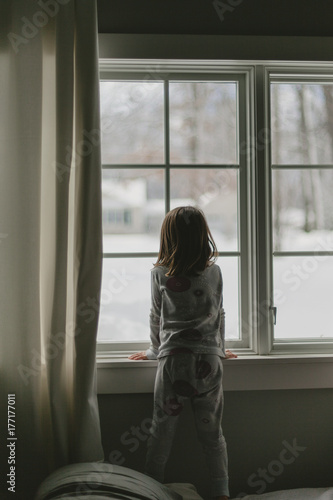 Girl in pajamas looking out window at snowy winter scene photo