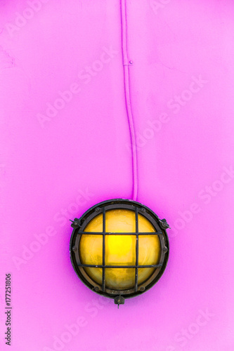 Ship yellow deck lamp (bulkhead light) surrounded by a metal rusted frame fixed to a painted light pink wooden wall and similarly pastel pink color painted electric cable.