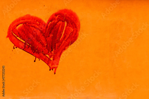 Red colored heart manually painted with drips in an orange wall with rustic rough imperfect surface.