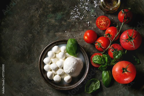 Ingredients for italian caprese salad. Mozzarella balls, buffalo in metal vintage plate, tomatoes, basil leaves, olive oil with vinegar over dark background. Top view with space. Rustic style photo