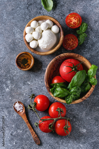 Ingredients for italian caprese salad. Mozzarella balls, buffalo, tomatoes, basil leaves, olive oil with vinegar, salt in olive wood bowls over gray texture background. Top view with copy space