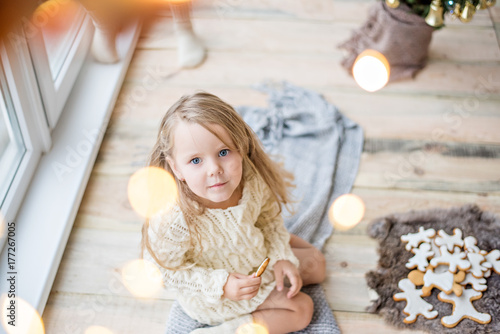 A little girl in a white knitted sweater drinks hot chocolate, cocoa. The baby looks out the window, waits for a New Year's miracle Santa Claus, eats Christmas gingerbread men with marshmallows. Card