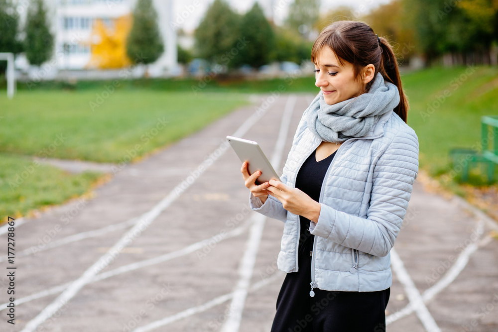 Young brunette sporty woman wiith tablet in hands dressed warm clothes and standing over stadium track background. Sport, lifestyle and multitacking concept. Young brunette sporty woman wiith tablet in hands dressed warm clothes and standing over stadium track background. Sport, lifestyle and multitacking concept.