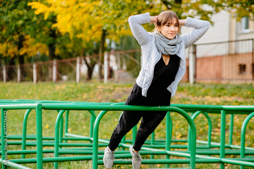 A young caucasian athlete girl in warm gray sportswear trains push-ups in an emphasis lying on the parallel bars of an sports gym for outdoor training. Autumn sports and healthy lifestyle