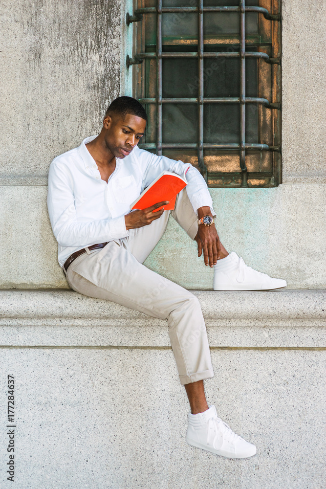 Young African American College Student studying in New York, wearing white shirt, light gray pants, sneakers, wristwatch, sitting against vintage wall with window on street, reading red book..