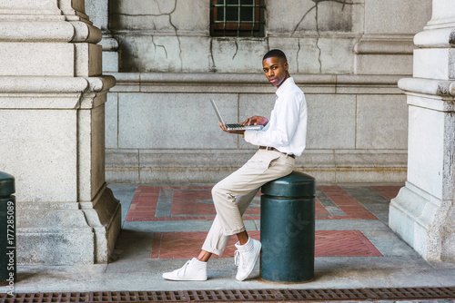 Young African American College Student studying New York, wearing white shirt, light gray pants, white sneakers, sitting on metal pillar on vintage street, working on laptop computer, looking away..