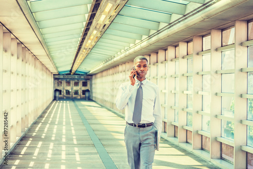 Young African American Businessman working in New York, wearing white shirt, necktie, holding laptop computer, walking on walkway with glass walls, ceiling, wooden floor, talking on cell phone..
