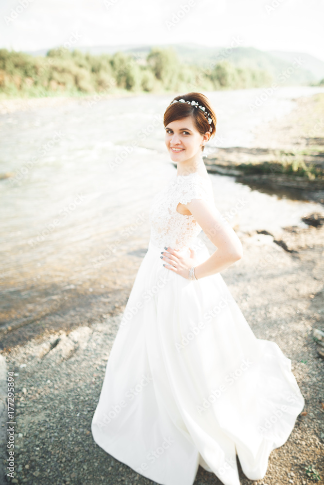 Gorgeous beautiful bride smiles while posing by the river with bouquet