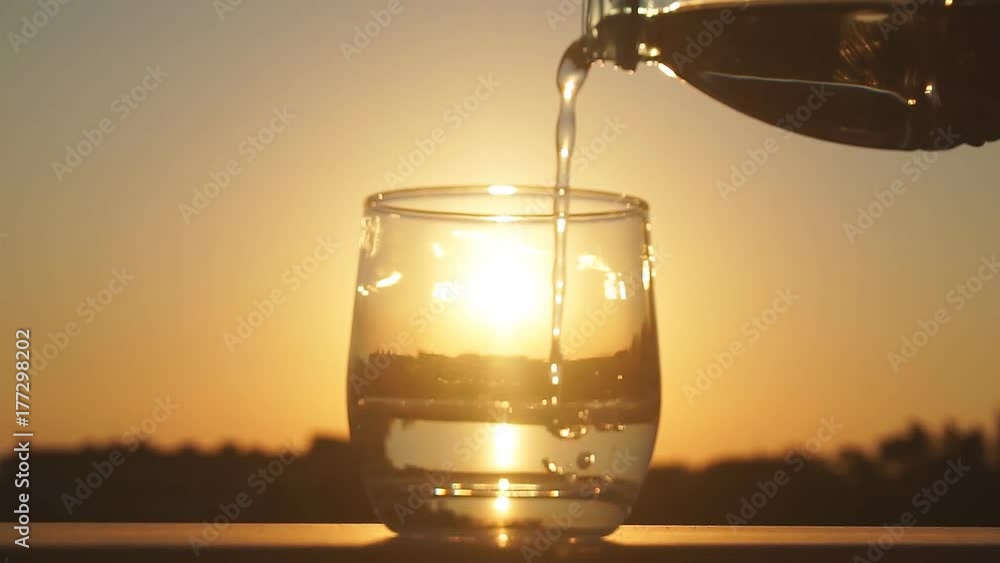 Slow motion close-up shot of pouring mineral water into glass on the background of big golden evening sun reflecting in the glass Slow motion close-up shot of pouring mineral water into glass on the background of big golden evening sun reflecting in the glass