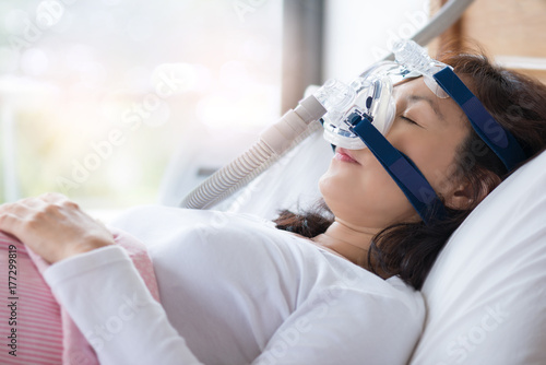 Woman and cpap mask, healthcare concept.Senior woman using cpap machine to stop choking and snoring from obstructive sleep apnea with bokeh and morning light background.. © sbw19