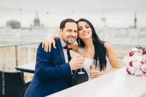 Cheerful married couple embrace each other, clink glasses with champagne, have good mood after registring their marriage, celebrate alone, wait for guests, pose at camera with pleased smiles photo