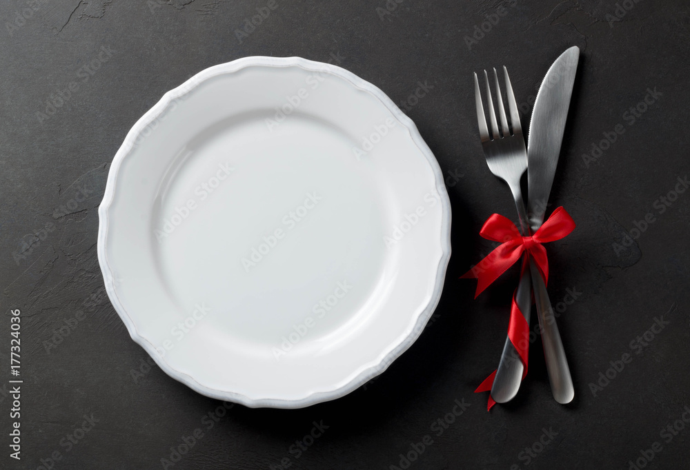 Festive set of cutlery knife and fork with red satin bow with a white plate, dark stone slate background, top view, copyspace