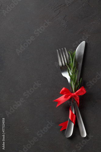 Festive set of cutlery knife and fork with red satin bow with rosemary, dark stone slate background, top view, copyspace, vertical image