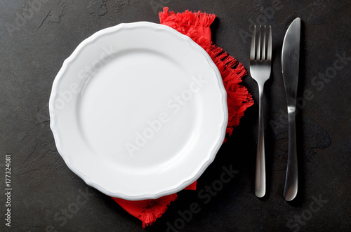 Festive set of knife and fork and white plate on a red napkin on a dark stone slate background, top view, copyspace