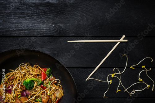 Udon stir fry noodles with chicken and vegetables in wok pan on black wooden background. With chopsticks