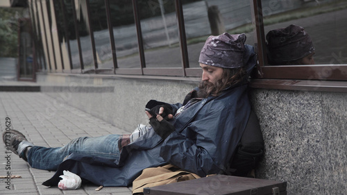 Bearded unemployed begging man sitting in street while using smartphone