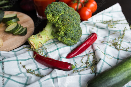  inflorescence of broccoli, a green squash and two red chili peppers are lying on the plaid tissue. in the background there are red tomatoes and a glass of tomato juice. Hurbs are used as decoration.