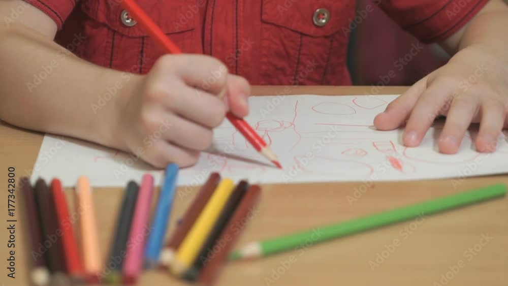 Close-up of hands of unidentified little boy dressed in red shirt sitting at table drawing pictures using multicolored pencils in kindergarten Close-up of hands of unidentified little boy dressed in red shirt sitting at table drawing pictures using multicolored pencils in kindergarten