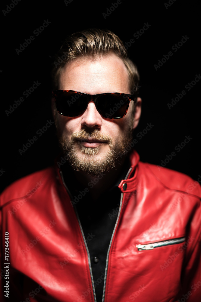 Cool man in a red leather jacket with sunglasses standing against a black background.